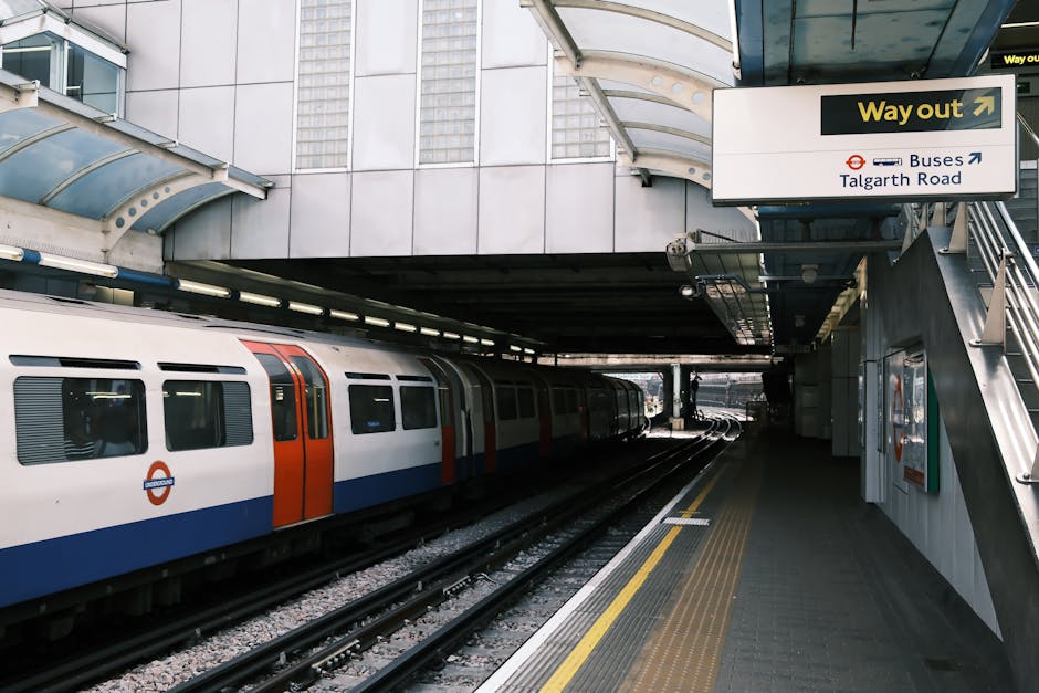 Exterior view of a London underground train station platform with a white, blue, and orange train on the tracks. The platform has a textured, non-slip surface with yellow tactile paving near the edge 