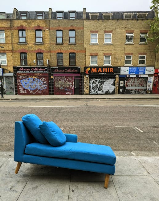An outdoor street scene showing a single blue upholstered chaise lounge with a matching cushion placed on a concrete sidewalk. The chaise features wooden legs and is positioned near the curb, with a row of closed commercial shopfronts with metal shutters covered in graffiti in the background. The buildings above have multiple windows and are constructed of brick with a weathered appearance. Natural daylight illuminates the scene, highlighting the clean, bright surface of the upholstery against the gritty urban setting. The image emphasizes surface cleaning and maintenance as provided by Hammersmith Carpet Cleaners, showcasing an outdoor furniture piece in a public space, illustrating the importance of deep cleaning and sanitisation for domestic and commercial environments.