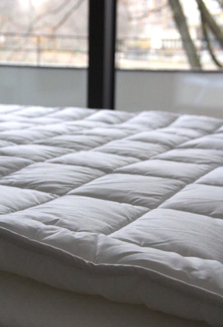 Close-up view of a freshly cleaned and sanitised white mattress with quilted stitching on a modern bed frame in a well-lit bedroom. The mattress surface appears smooth, clean, and free of dust or stains, indicating recent deep cleaning. In the background, large windows with natural daylight streaming in reveal an outdoor view with trees and building structures, emphasizing a bright and airy atmosphere. The room’s surfaces and fabrics show no visible dirt or clutter, reflecting professional domestic cleaning by Hammersmith Carpet Cleaners. The focus highlights surface cleaning and sanitisation processes aimed at maintaining hygiene for residents, including those with children and pets, as promoted on the Ravenscourt Park mattress cleaning page for Hammersmith.